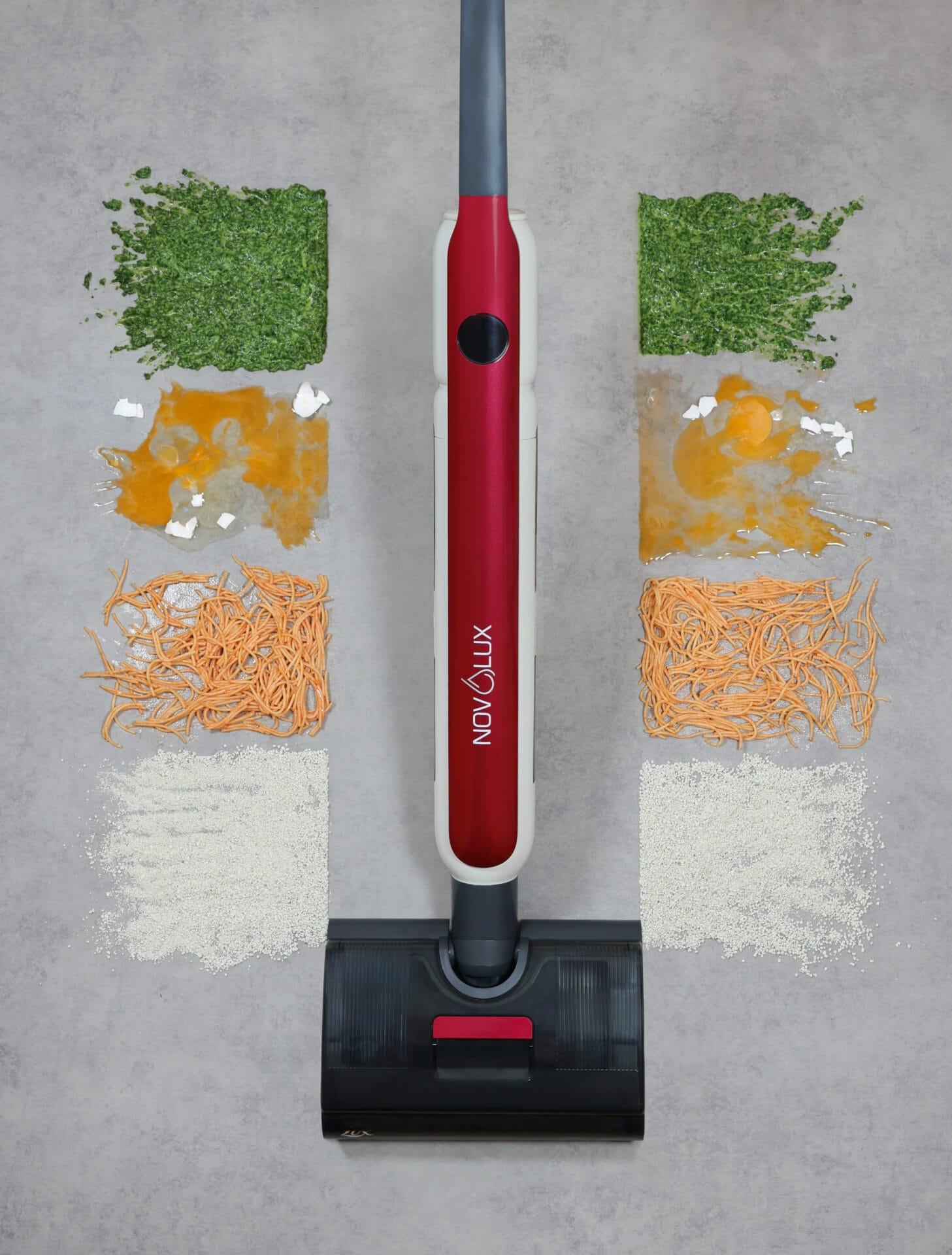Ein rot-schwarzer Stabstaubsauger steht aufrecht auf einer grauen Fläche, auf der verschiedene Essensreste - darunter Kräuter, Eigelb, Nudeln und Körner - in ordentlichen, bunten Quadraten auf beiden Seiten des Staubsaugerwegs angeordnet sind.